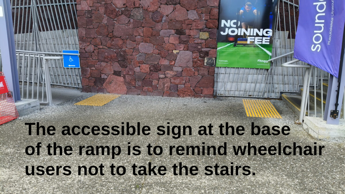 The entrance to the Mt Albert Community Centre, Auckland. To the left a ramp with an accessible sign, to the right stairs. Text: The accessible sign at the base of the ramp is to remind wheelchair users not to take the stairs.